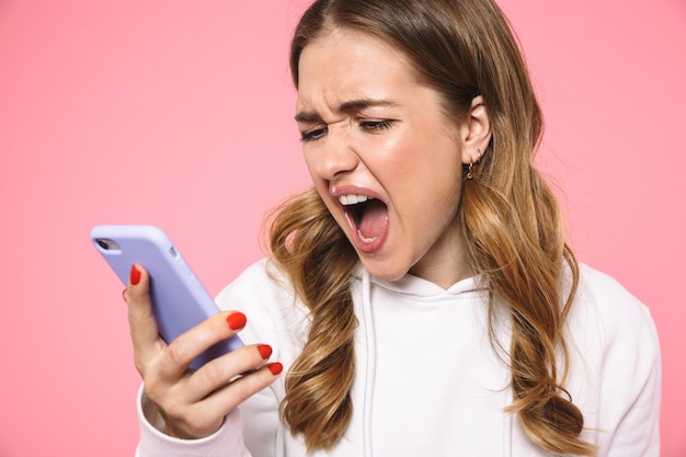 Foto mulher loira gritando usando roupas casuais falando pelo smartphone na parede rosa