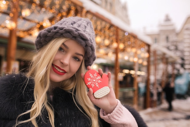 Mulher loira incrível segurando um saboroso pão de gengibre contra a decoração leve na feira de Natal em Kiev