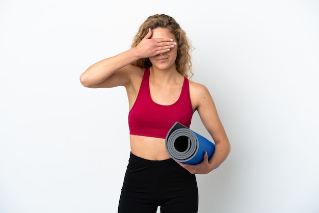 Mulher loira jovem esporte indo para aulas de ioga, segurando um tapete isolado no fundo branco, cobrindo os olhos com as mãos. Não quero ver nada