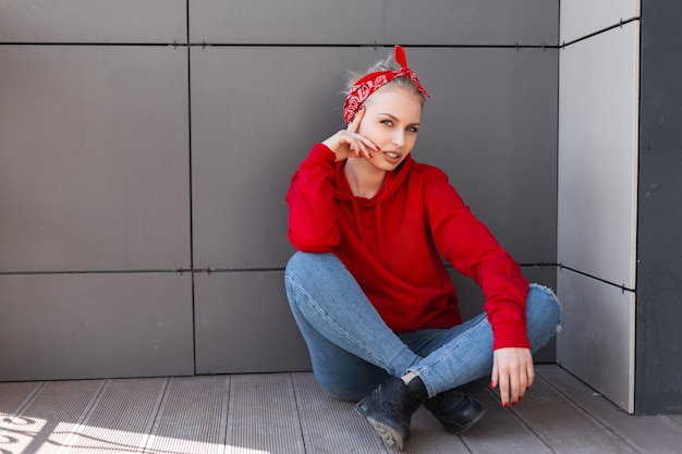 Mulher loira jovem muito feliz em uma bandana elegante com um padrão em jeans azul em um moletom vermelho da moda com botas pretas, descansando sentada perto de uma parede cinza em um dia de verão