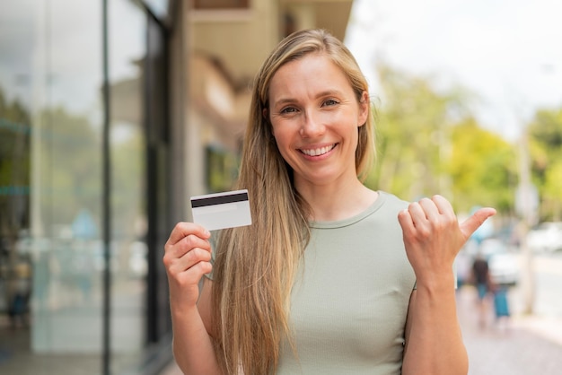 Mulher loira jovem segurando um cartão de crédito ao ar livre apontando para o lado para apresentar um produto