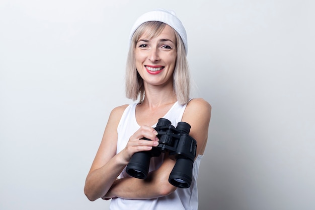 Mulher loira jovem sorridente com binóculos sobre um fundo claro.