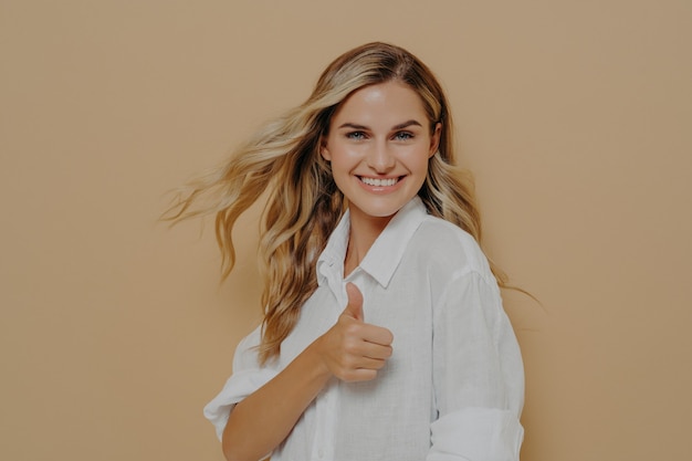 Mulher loira linda animada com cabelo longo ondulado esvoaçante posando com o polegar para cima sinal de ok, gesticulando que tudo está ok com o dedo para cima, isolado sobre o fundo bege do estúdio com espaço de cópia