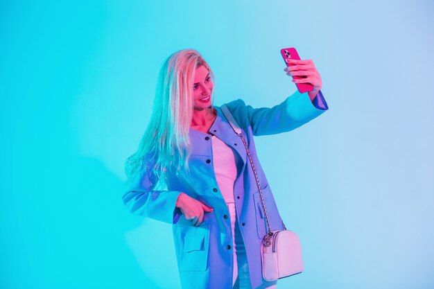 Mulher loira linda feliz com um sorriso fofo em roupas de negócios da moda olha com uma bolsa e um blazer faz uma foto de selfie no telefone inteligente no estúdio em uma luz multicolorida