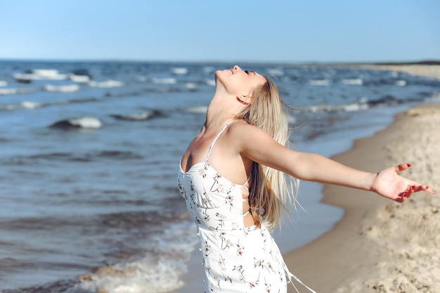 Mulher loira linda feliz na praia do oceano em pé com um vestido branco de verão, levantando as mãos.