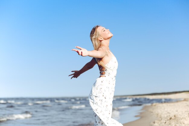Mulher loira linda feliz na praia do oceano em pé com um vestido branco de verão, levantando as mãos.
