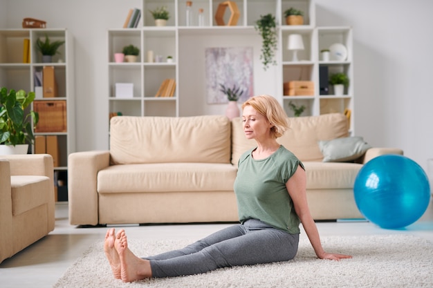 Mulher loira madura e ativa em roupas esportivas, sentada no chão e se exercitando na sala de estar enquanto fica em casa durante a quarentena