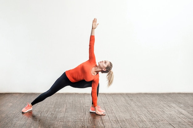 Mulher loira magro em roupas esportivas apertadas, praticando ioga, em pé na pose do triângulo trikonasana, treinando os músculos para flexibilidade. Saúde, atividade esportiva e ginástica em casa. foto de estúdio interno