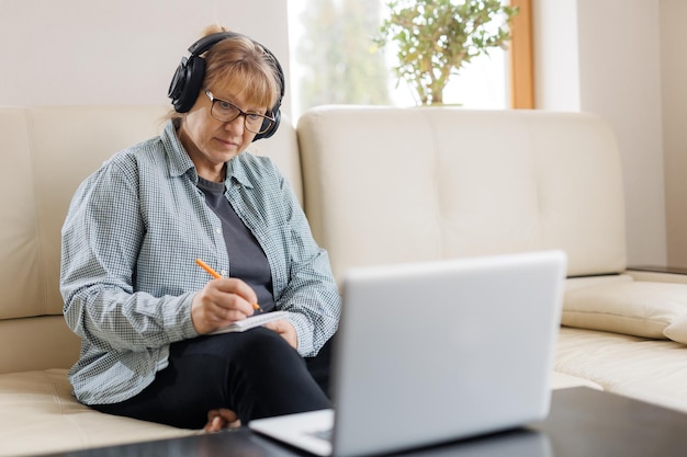 Mulher loira moderna de meia-idade em fones de ouvido faz curso on-line ou treinamento em computador em casa Mulher caucasiana madura inteligente em fones de ouvido assiste a webinar faz anotações estuda distante no laptop