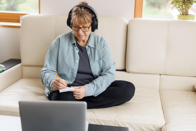 Mulher loira moderna de meia-idade em fones de ouvido faz curso on-line ou treinamento em computador em casa Mulher caucasiana madura inteligente em fones de ouvido assiste a webinar faz anotações estuda distante no laptop