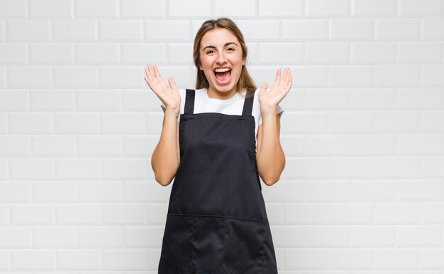 Mulher loira parecendo feliz e animada, chocada com uma surpresa inesperada com as duas mãos abertas ao lado do rosto