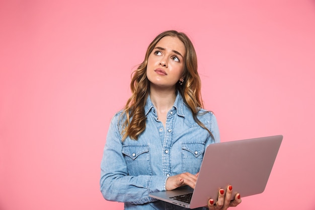 Mulher loira pensativa, vestindo uma camisa jeans, usando um computador laptop e olhando por cima da parede rosa