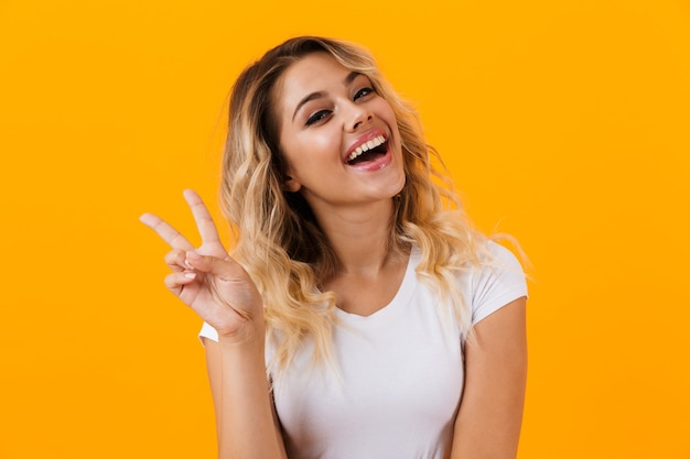 Foto mulher loira positiva com roupas básicas sorrindo e mostrando o símbolo da paz, isolada sobre a parede amarela