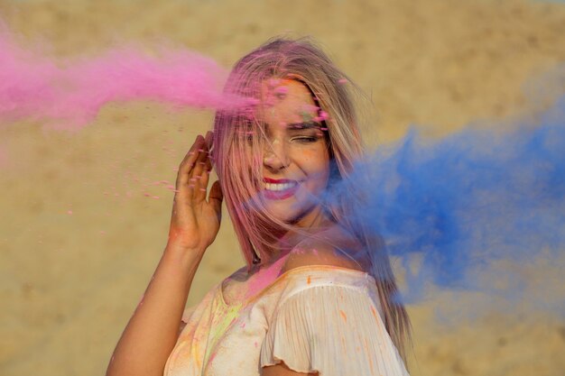 Mulher loira positiva posando em uma nuvem de tinta seca rosa e azul Holi no deserto