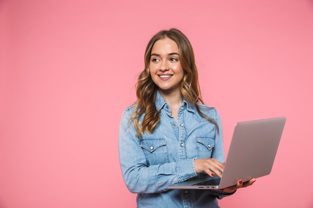 Mulher loira satisfeita vestindo uma camisa jeans, usando um computador laptop e olhando para longe por cima da parede rosa