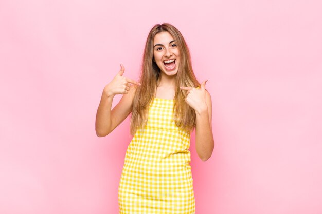 Mulher loira se sentindo feliz, surpresa e orgulhosa, apontando para si mesma com um olhar animado e surpreso