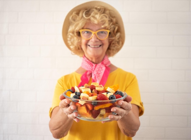 Mulher loira sênior feliz desfocada com chapéu isolado em um fundo branco detém uma tigela com um conceito de alimentação saudável de salada de frutas frescas e deliciosas de verão