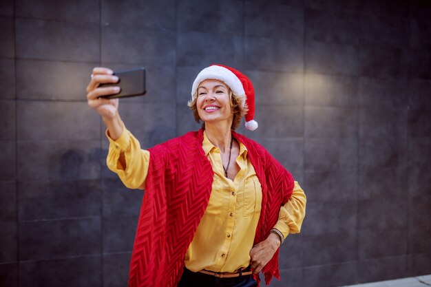 Mulher loira sênior sorridente atraente com chapéu de Papai Noel na cabeça, posando e tomando selfie em frente a parede ao ar livre.