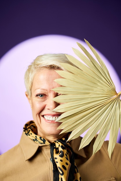 Mulher loira sênior sorridente com cabelo curto se escondendo atrás do ventilador bege