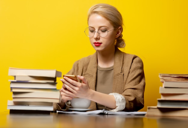 Mulher loira sentada à mesa com livros e usando o celular em amarelo