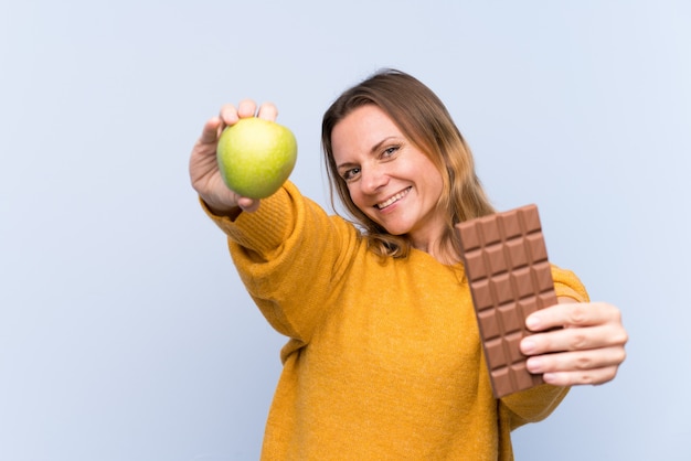 Mulher loira sobre parede azul isolada, tomando uma tablete de chocolate em uma mão e uma maçã na outra