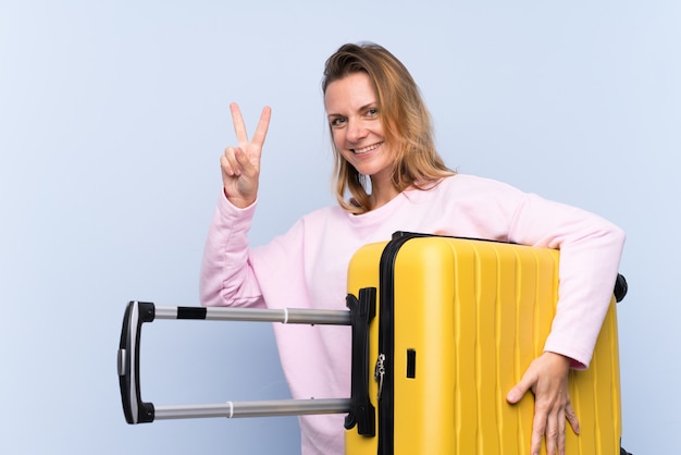 Mulher loira sobre parede isolada em férias com mala de viagem e fazendo o gesto de vitória