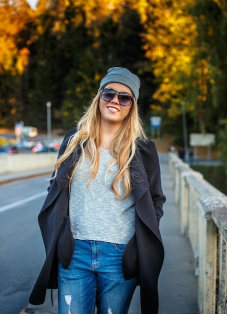 Mulher loira sorridente em óculos de sol e roupas casuais posando sobre a floresta de outono.