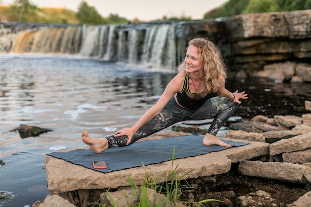Mulher loira sorridente em roupas esportivas, praticando ioga à beira de um rio em frente a uma cachoeira.