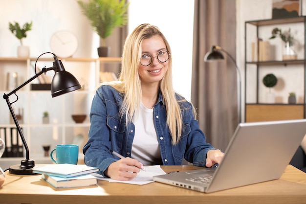 Mulher loira sorridente escrevendo notas enquanto estiver usando o computador portátil sentado na mesa em casa