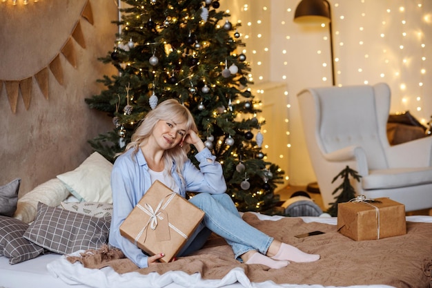 Mulher loira sorridente sentada na cama com um presente de Natal em frente à árvore de Natal e as luzes