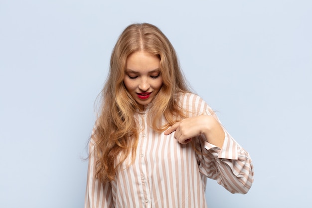 Mulher loira sorrindo alegre e casualmente, olhando para baixo e apontando para o peito