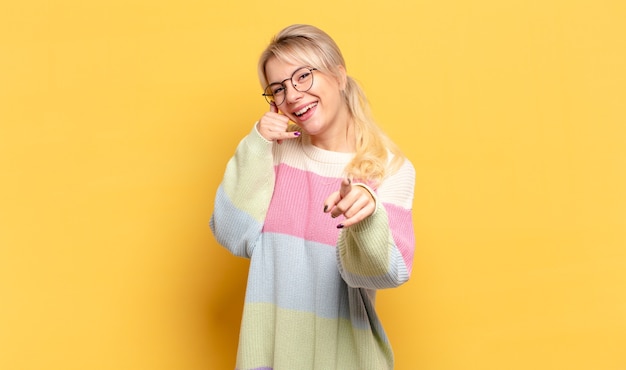 Mulher loira sorrindo alegremente e apontando para a câmera enquanto faz um gesto depois de ligar para você, falando ao telefone