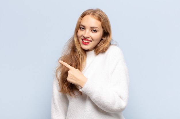 Mulher loira sorrindo alegremente, sentindo-se feliz e apontando para o lado e para cima, mostrando o objeto no espaço da cópia