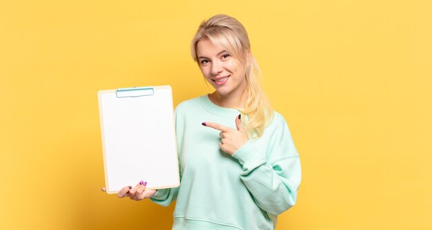 Mulher loira sorrindo alegremente, sentindo-se feliz e apontando para o lado e para cima, mostrando o objeto no espaço da cópia