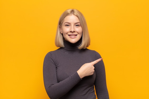 Mulher loira sorrindo alegremente, sentindo-se feliz e apontando para o lado e para cima, mostrando o objeto no espaço da cópia