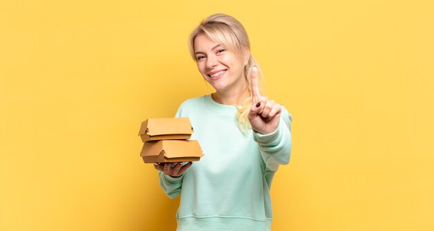 Mulher loira sorrindo com orgulho e confiança fazendo a pose número um triunfantemente, sentindo-se uma líder