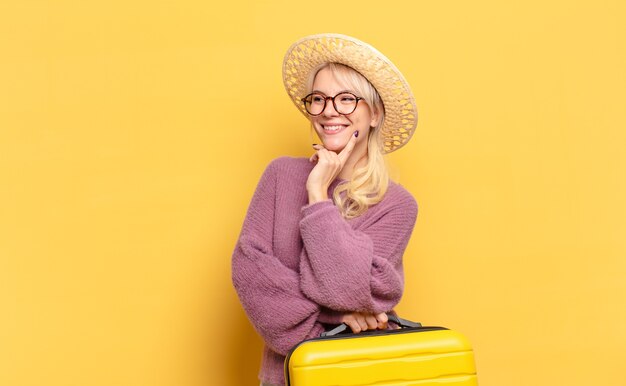 Mulher loira sorrindo com uma expressão feliz e confiante com a mão no queixo, pensando e olhando para o lado