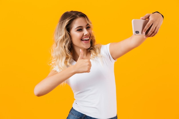 Mulher loira sorrindo e tirando foto de selfie no celular, isolada na parede amarela