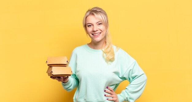 Mulher loira sorrindo feliz com uma mão no quadril e atitude confiante, positiva, orgulhosa e amigável