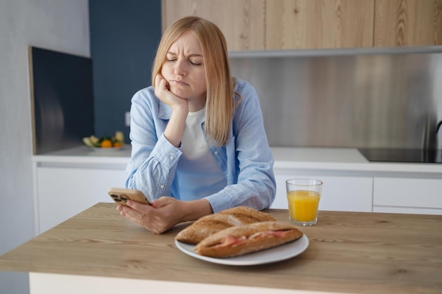 Mulher loira triste lendo mensagem ou má notícia no smartphone na cozinha enquanto cozinha o café da manhã em