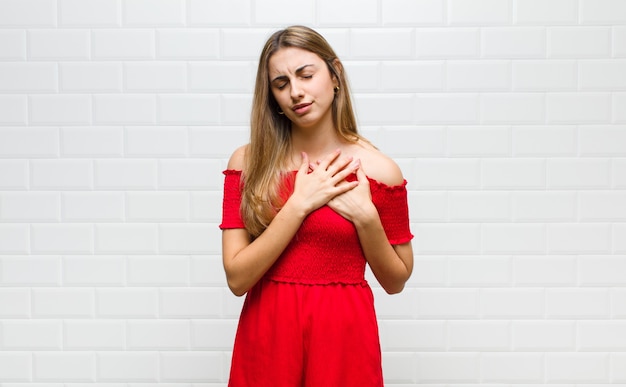 Mulher loira triste, magoada e com o coração partido, segurando as duas mãos perto do coração, chorando e se sentindo deprimida