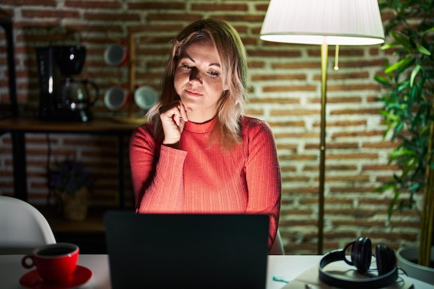 Mulher loira usando laptop à noite em casa com a mão no queixo pensando em pergunta expressão pensativa sorrindo com o conceito de dúvida de rosto pensativo
