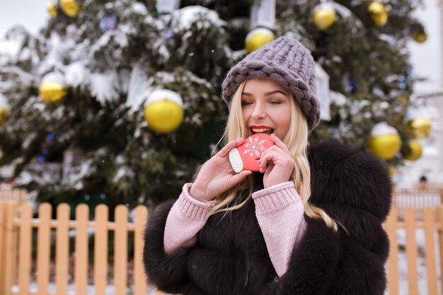 Mulher loira vestida com estilo e chapéu de malha cinza morde um delicioso pão de gengibre contra uma árvore de Natal na cidade