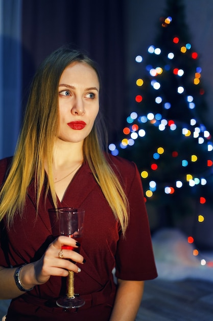 Mulher loira vestida de vermelho com uma taça de vinho branco ou champanhe em casa. Árvore de Natal. Decorações de ano novo