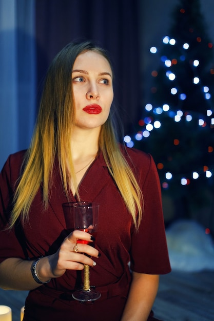 Mulher loira vestida de vermelho com uma taça de vinho branco ou champanhe em casa. Árvore de Natal. Decorações de ano novo