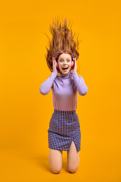 Mulher louca alegre emocional em fones de ouvido com cabelo a voar isolado no fundo de cor amarela em estúdio, retrato. Mulher caucasiana animada com cabelo ruivo natural na camisa e saia curtir música