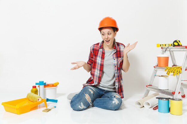 Mulher louca divertida no capacete protetor laranja, sentado no chão com instrumentos para apartamento de renovação isolado no fundo branco. Papel de parede, acessórios para colar, ferramentas de pintura. Conceito de reparo.