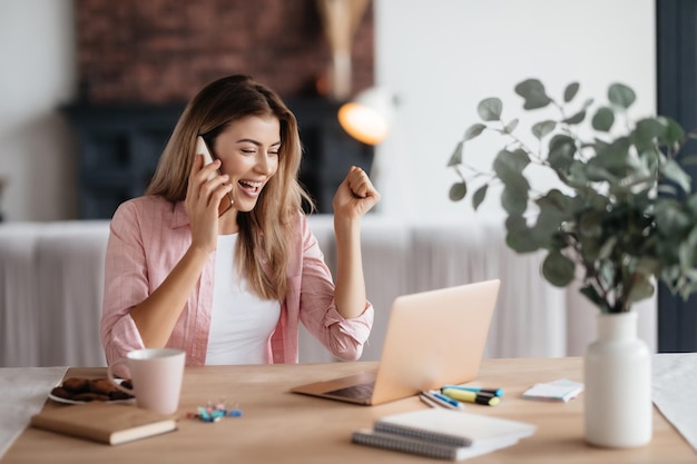 Mulher loura animada sorrindo amplamente enquanto faz um telefonema e levanta a mão triunfante