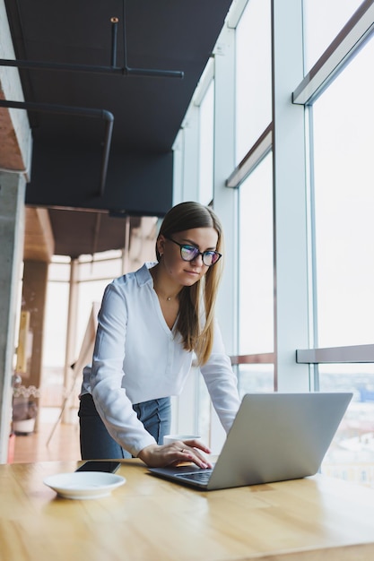 Mulher loura atraente em roupas casuais e óculos está se concentrando e digitando em um laptop enquanto está sentado em uma mesa em um café com café uma estudante em uma camisa branca e óculos