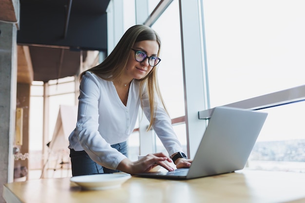 Mulher loura atraente em roupas casuais e óculos está se concentrando e digitando em um laptop enquanto está sentado em uma mesa em um café com café uma estudante em uma camisa branca e óculos